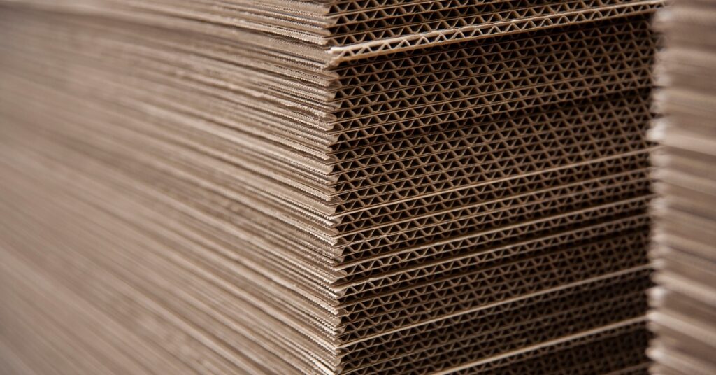 There are two long stacks of large, flat sheets of cardboard, displaying the side with the flutes within them.
