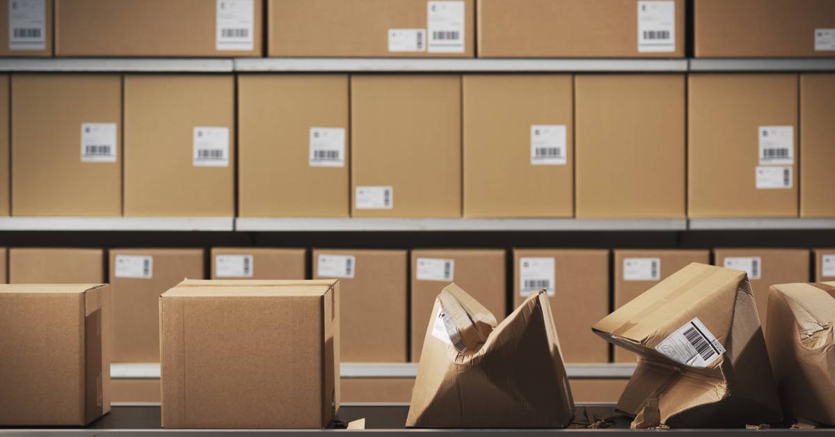 A warehouse with a wall of stacked boxes has a conveyor belt with boxes. Three of the boxes on the belt have been crushed.