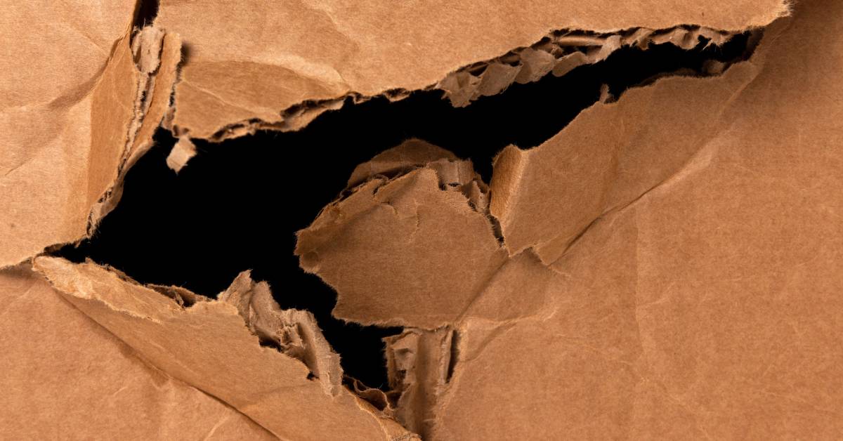 A close-up of a thin, brown cardboard box that has been ripped at one of the corners. The interior flutes are displayed.