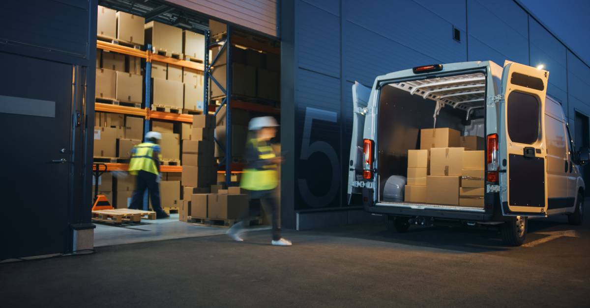 The back of a van has boxes inside of it, outside of an open building filled with stacked boxes. Employees are moving around.