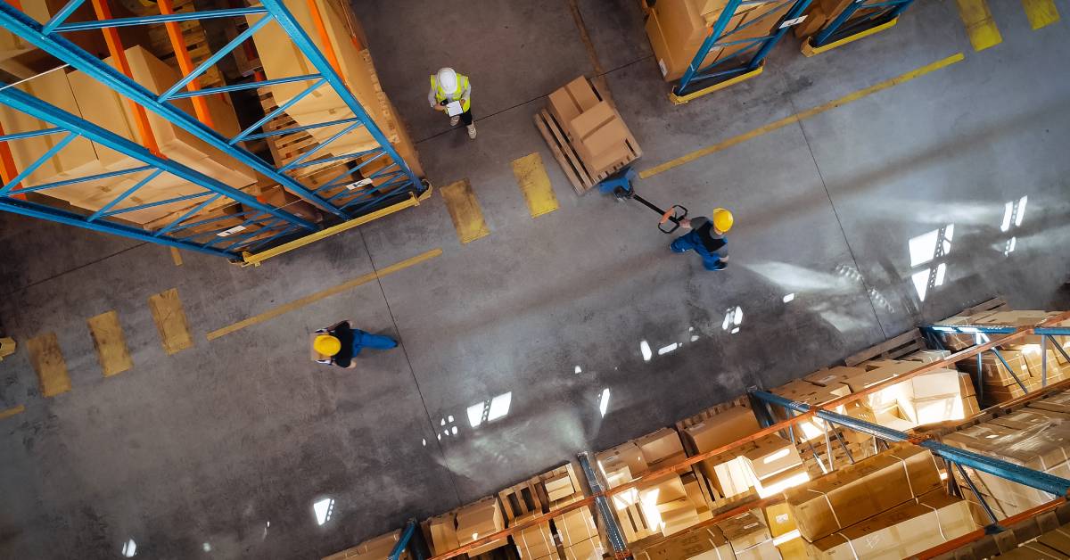 A top-down view of a warehouse with workers and many rows of stacked boxes on shelves. One worker has a dolly with boxes.