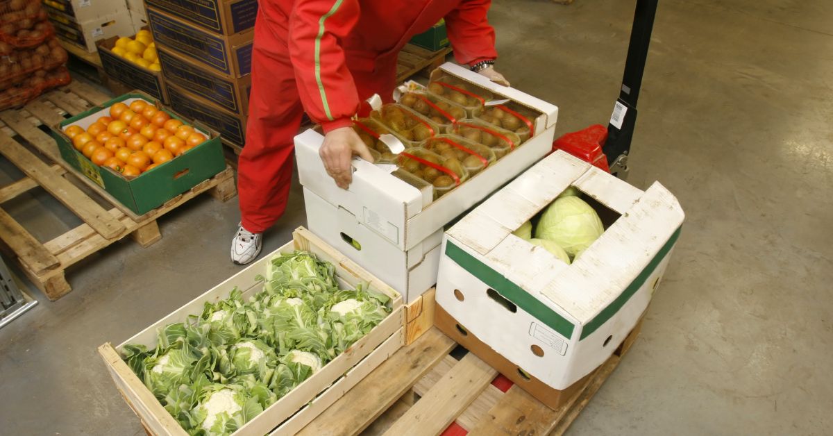 A man in a jumpsuit is unloading a dolly with boxes of cabbage and potatoes on it. A box of orange tomatoes are behind him.