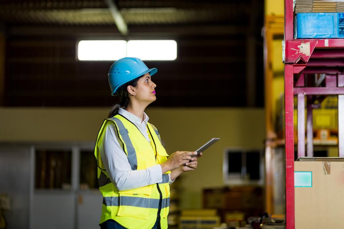 A worker wearing a hard hat and yellow vest is looking at boxes on a shelf in a warehouse. She is holding a tablet.