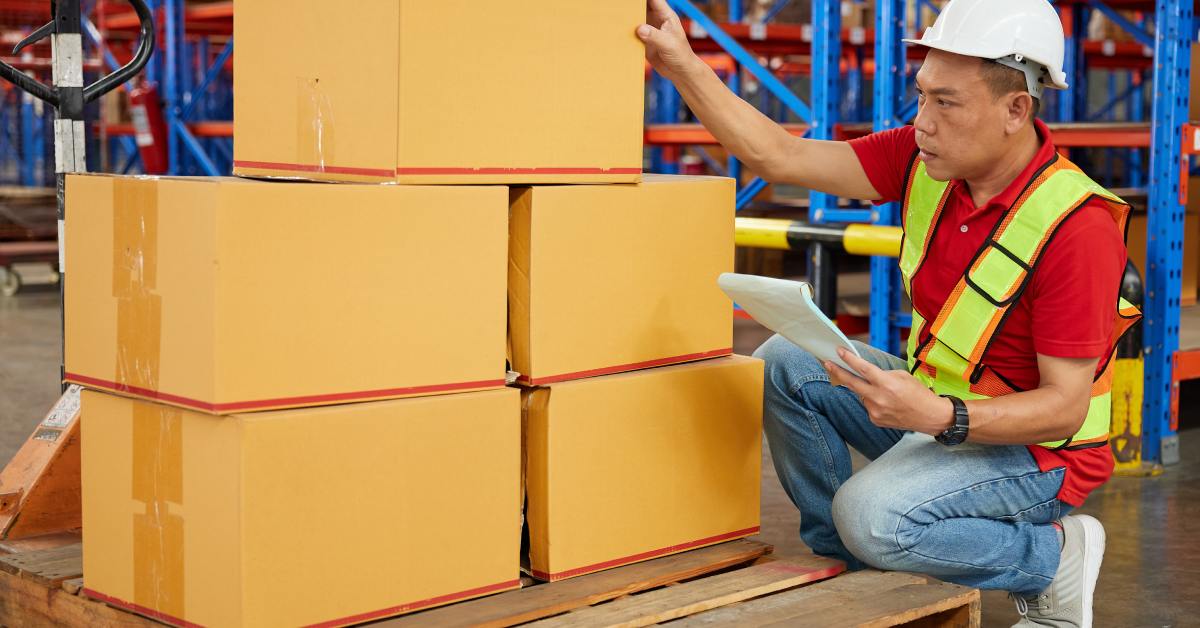 A man wearing a hard hat and yellow vest is crouching down next to a stack of boxes on a pallet in a warehouse.