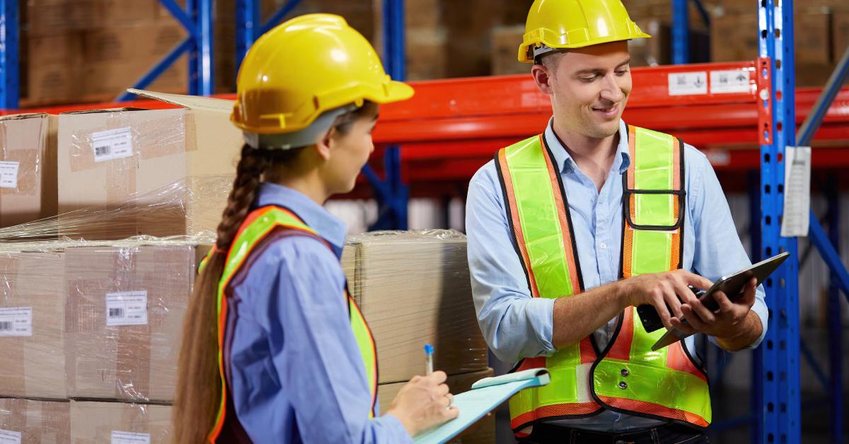 A man and woman wearing hard hats and yellow vests are talking next to a stack of boxes in a warehouse.