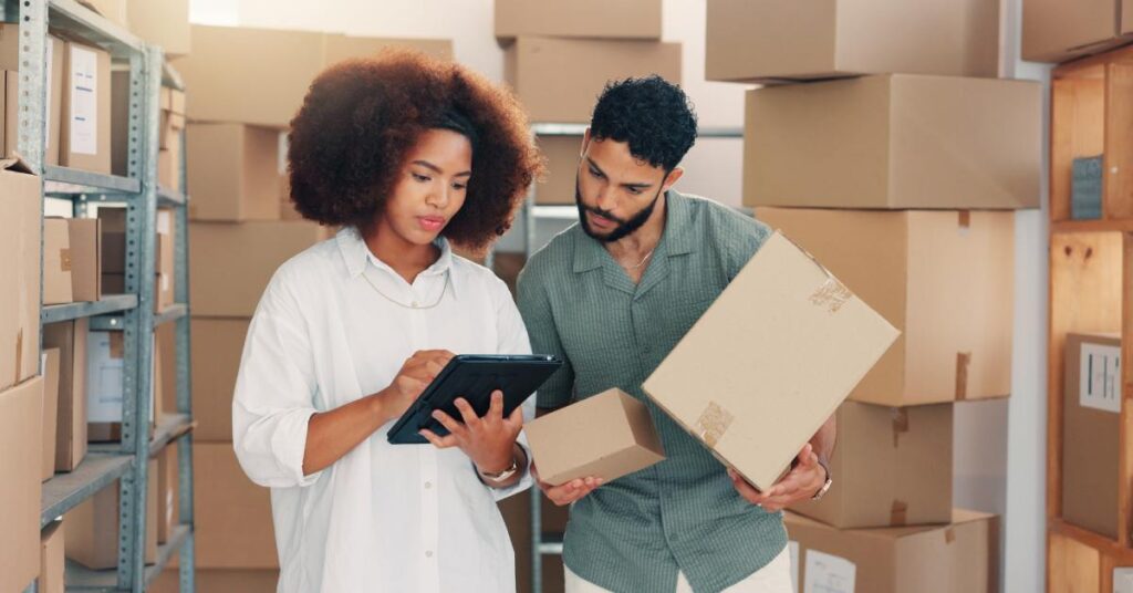 A man and woman are standing in a warehouse of boxes, looking at a device with a screen that the woman is holding.