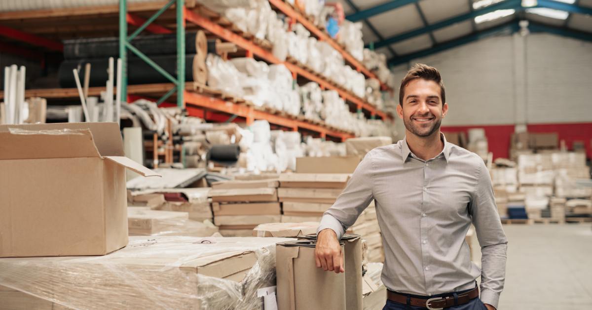 A smiling man is posing in professional attire in a warehouse of materials. He is leaning against an industrial roll of paper.