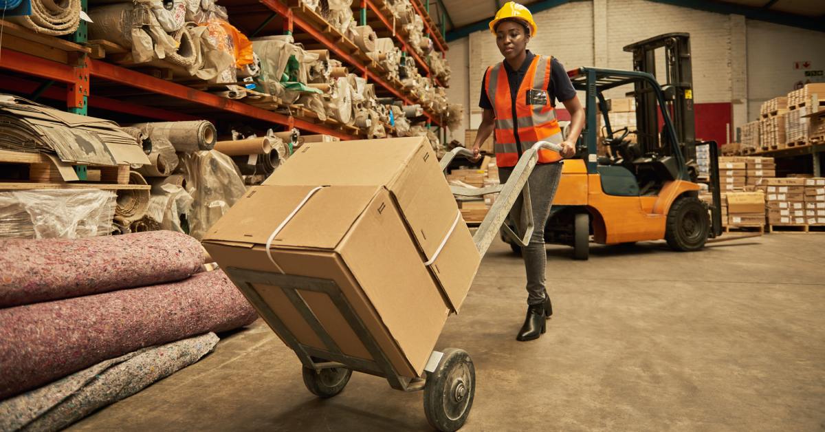 A female worker with a hard hat and vest is pushing a dolly with boxes in a warehouse. A forklift is behind her.