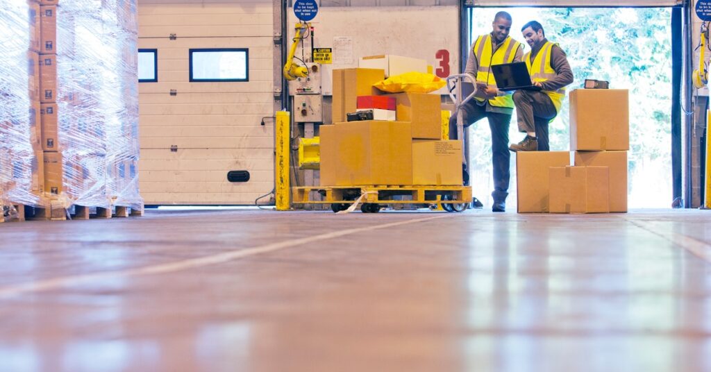 Two workers in a warehouse are standing alongside a stack of boxes on the ground and on a pallet while looking at a laptop.