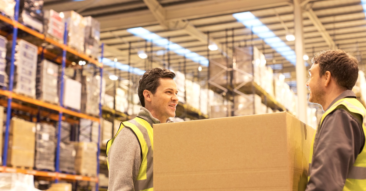 Two workers in a warehouse are holding two opposite ends of a large box. There are shelves of boxes in the background.