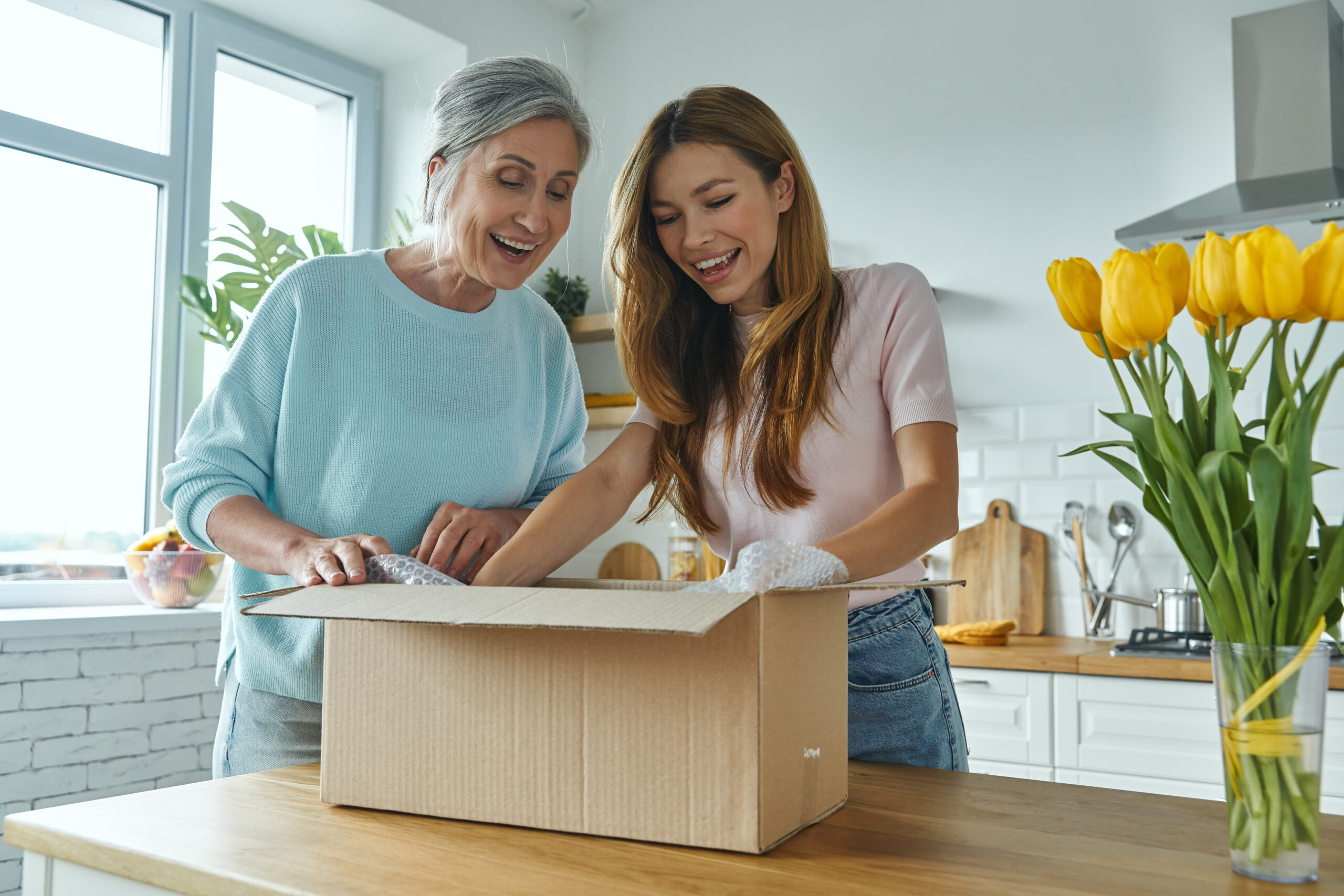 Two women are looking into a package. The package is sitting on a counter in a kitchen. There is a vase of flowers next to the package.