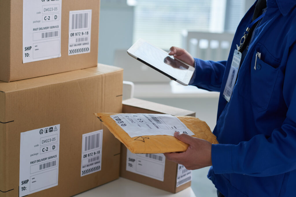 A person in a blue shirt is holding a tablet in their right hand and a package in their left. There are more packages next to them.