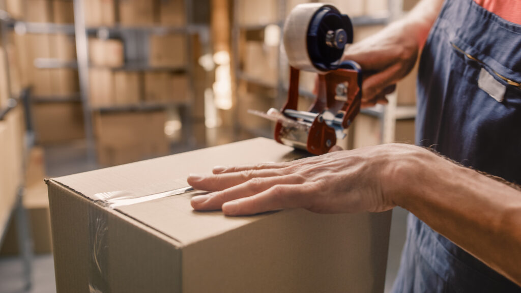 A person is taping a package shut. There are shelving units in the background, the shelves are filled with boxes.