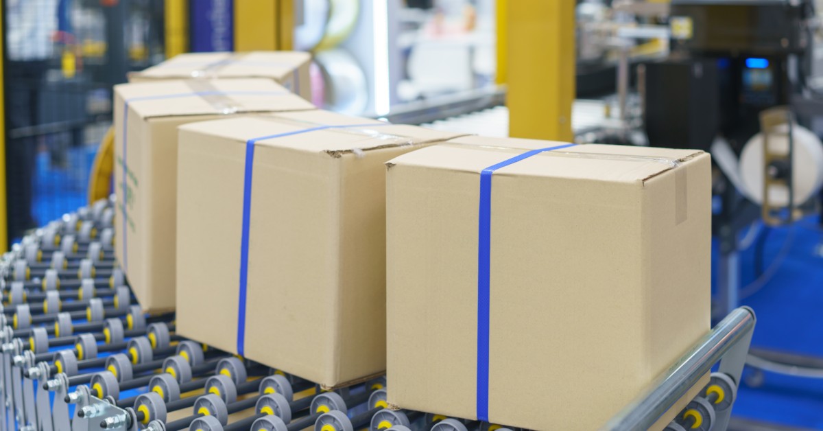 Four square boxes sit at the end of a conveyor belt in a shipping facility. Each box has blue tapped wrapped around it.