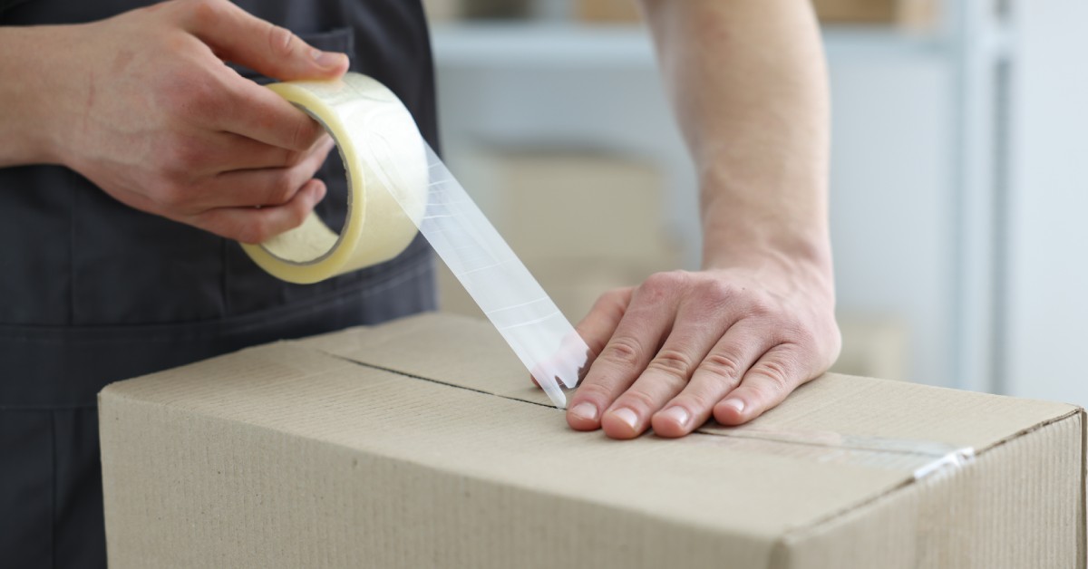 A person wearing a black shirt presses clear tape on a cardboard box to seal two of the flaps together.