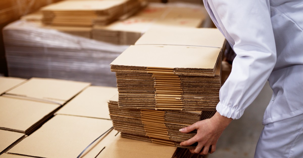 A factory worker is picking up a small stack of folded cardboard from a larger stack of cardboard.