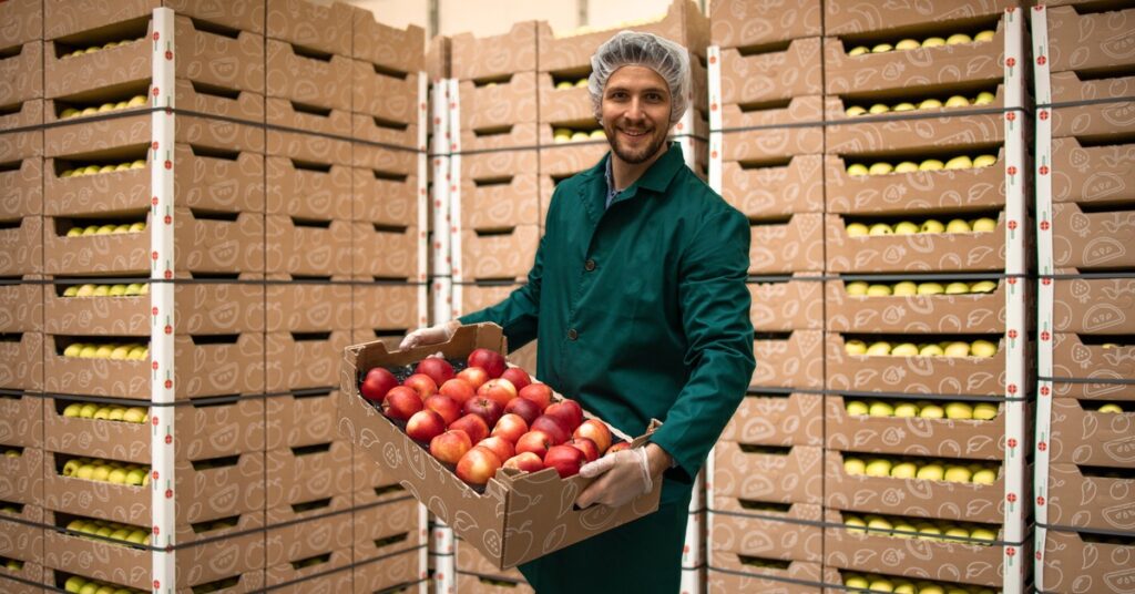 A man wearing a hairnet and coveralls is holding a box of produce. There are boxes behind him against the wall.