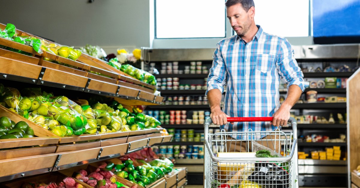 A man in a blue shirt is pushing a shopping cart. He is looking down at several bins of produce on a shelf.