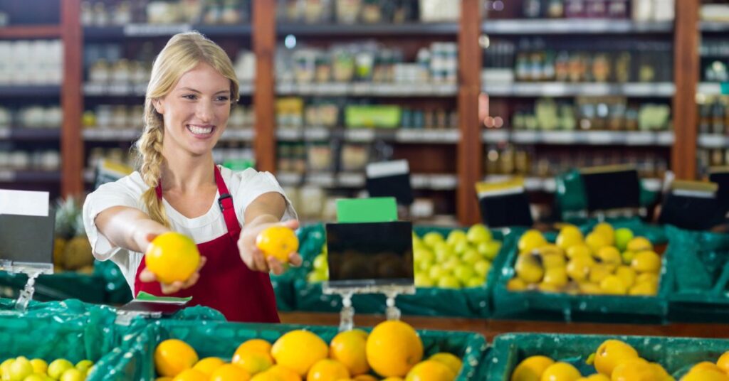A smiling woman in a red apron is holding fruit in her hands. There are several bins of fruit around her.