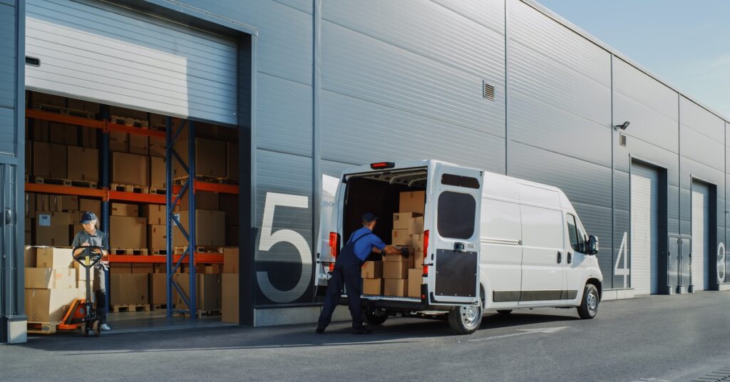 A man is loading boxes into the back of a white van. There is a warehouse next to him filled with more boxes.