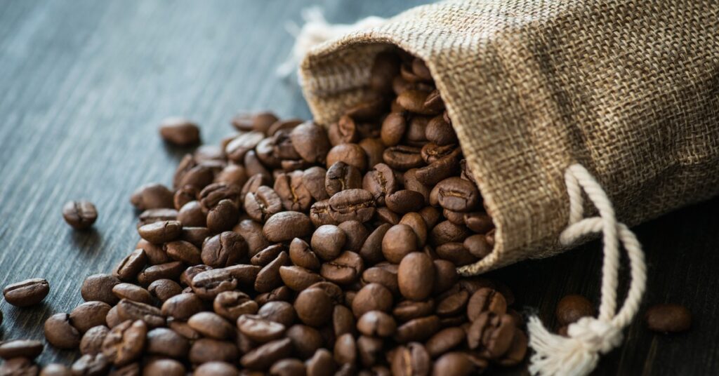 A closeup of coffee beans spilling out from a small burlap sack onto a wooden tabletop.