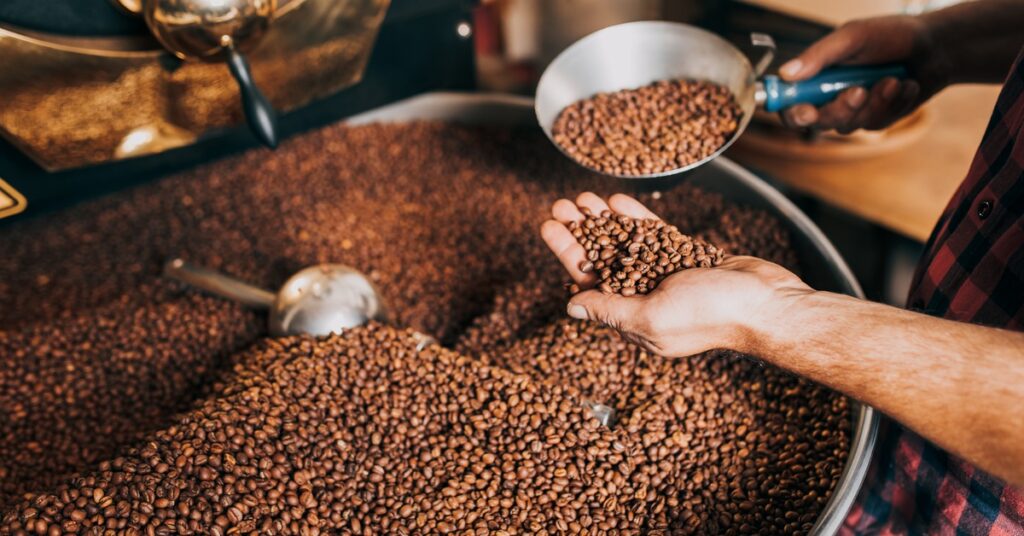 A man is holding out his hand and pouring coffee beans into his palm. Under his palm is a large pot of beans.