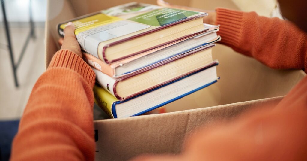A person is holding a stack of books. They are holding that stack above an open cardboard box.