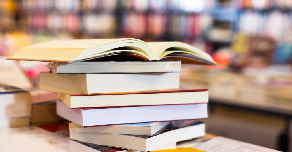 A stack of books is sitting on a table. In the background, there are shelves of books that run from one end of the frame to the other.