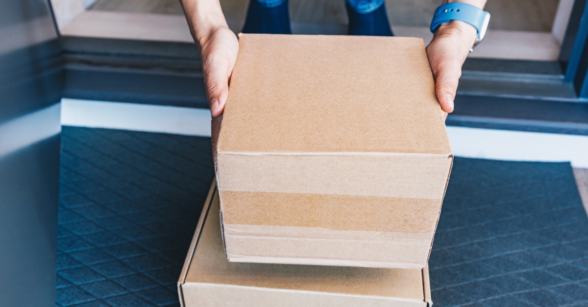 There are two boxes sitting on the ground. A person is bending over to pick up the box on top of the stack.
