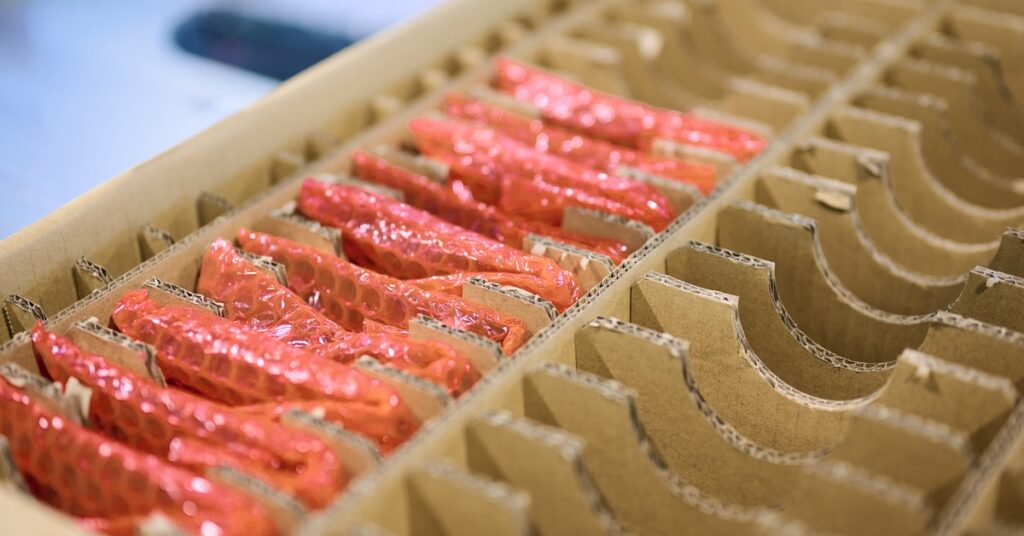 A cardboard box has several rows of slots. In some of the slots are items wrapped in bubble wrap.