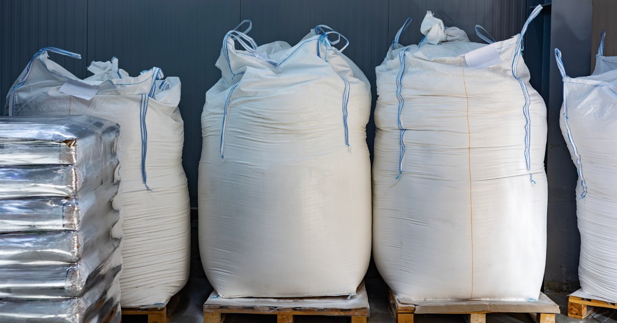 Several white bags are all on wooden pallets and lined up against a gray wall; all the bags have blue handles.