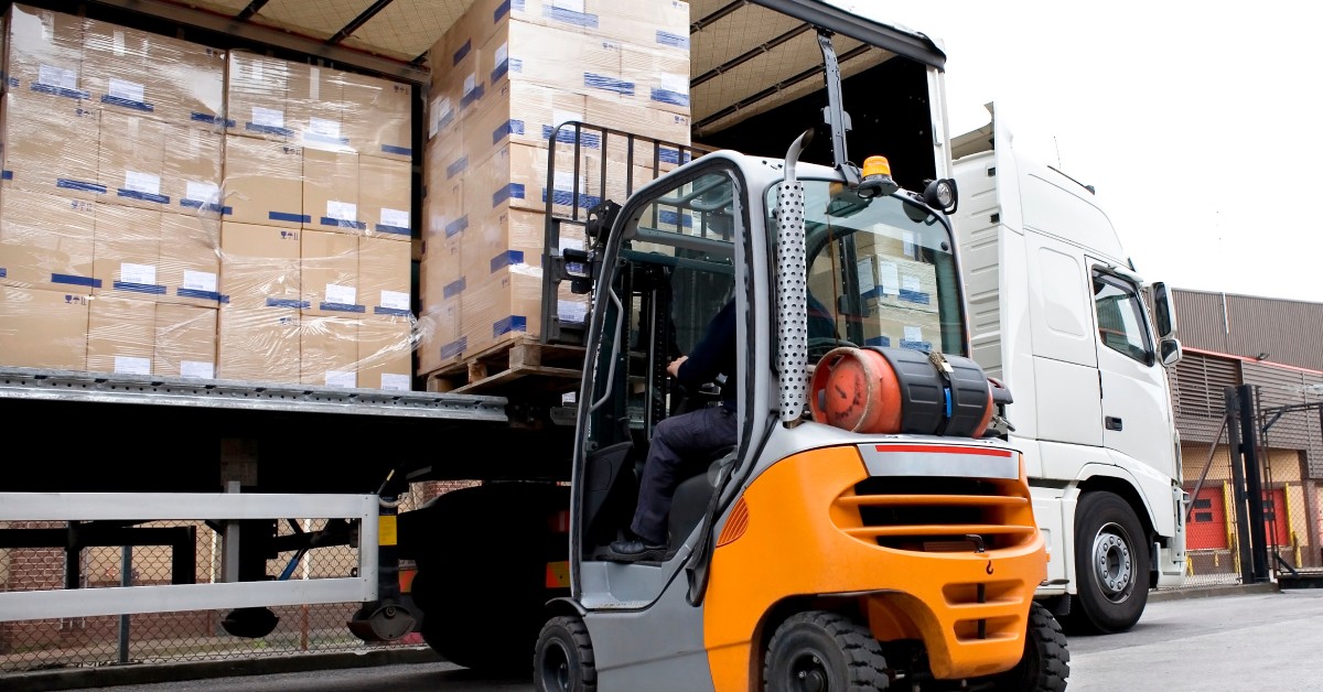 A person is in a forklift and is using it to take a pallet of boxes out of a truck trailer. The pallets are all stacked with boxes.