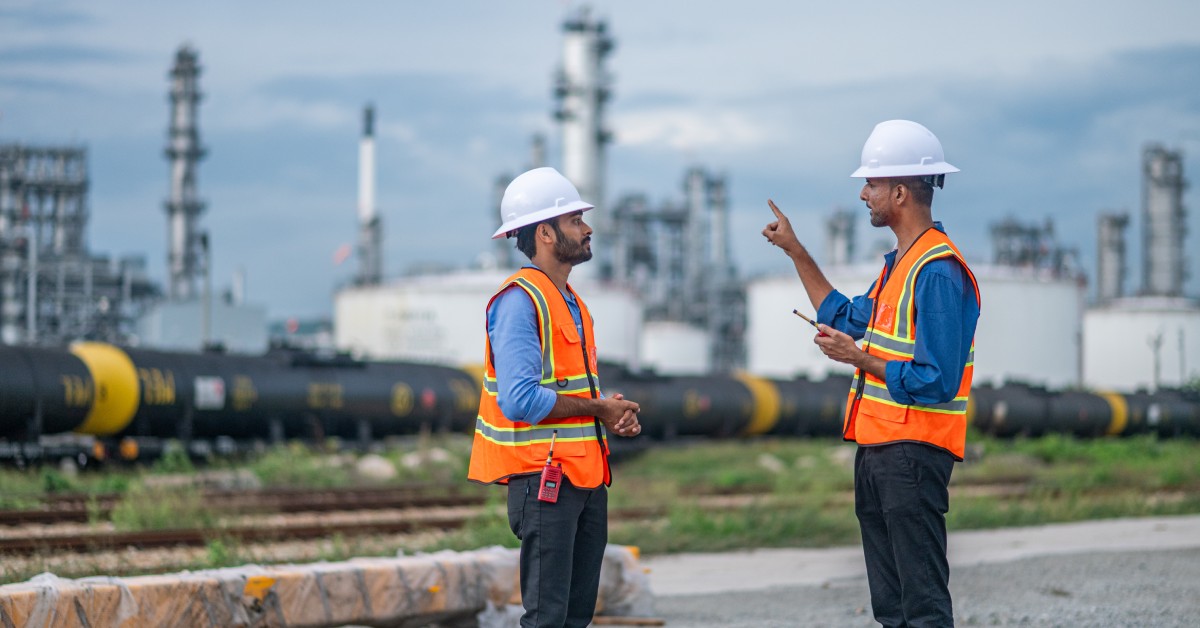 Two men, who are both wearing hard hats and orange vests, are standing outside and facing each other.