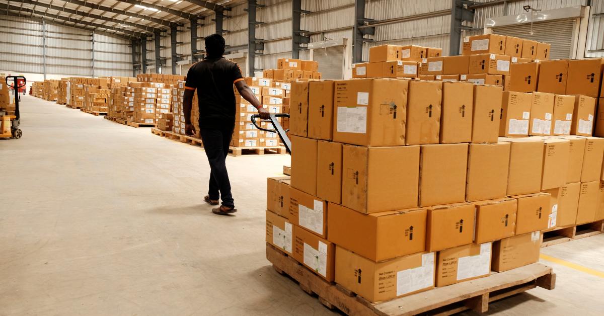 A man is pulling a pallet jack that has a pallet on it. There are stacks of boxes on the pallet that the man is pulling.