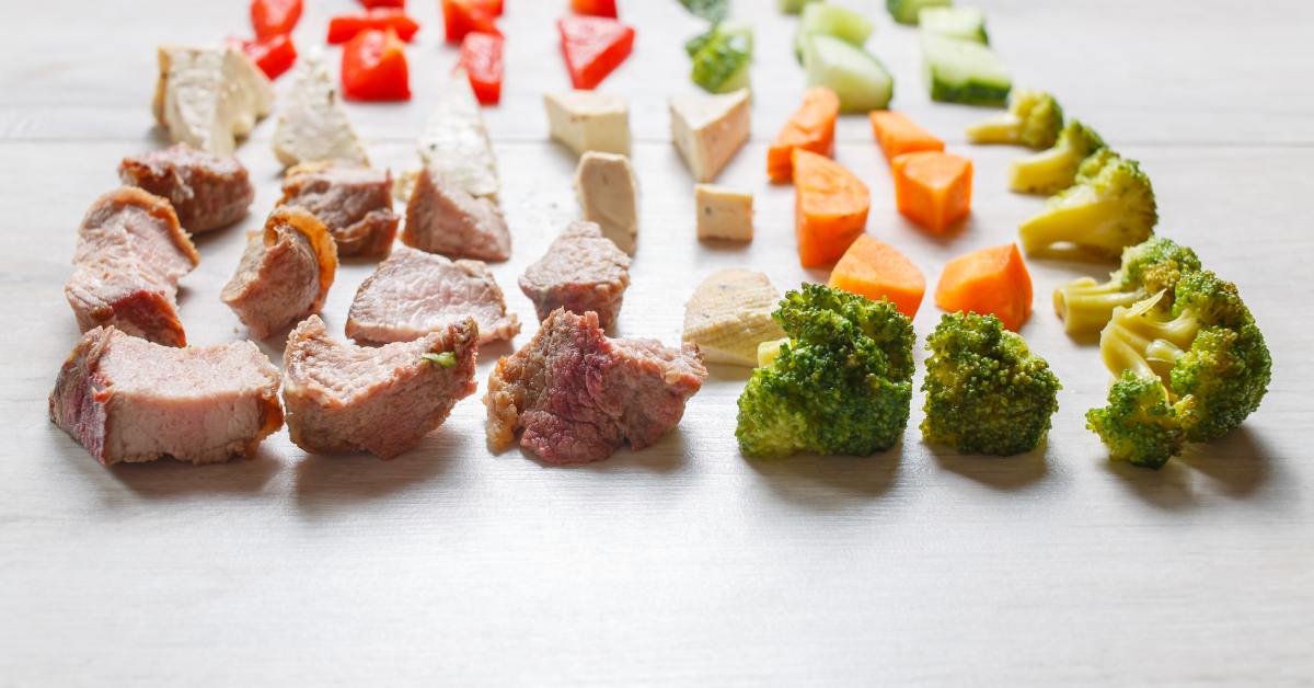 Different types of food are cut into pieces and lined up. There are pieces of broccoli, meat, and other vegetables.