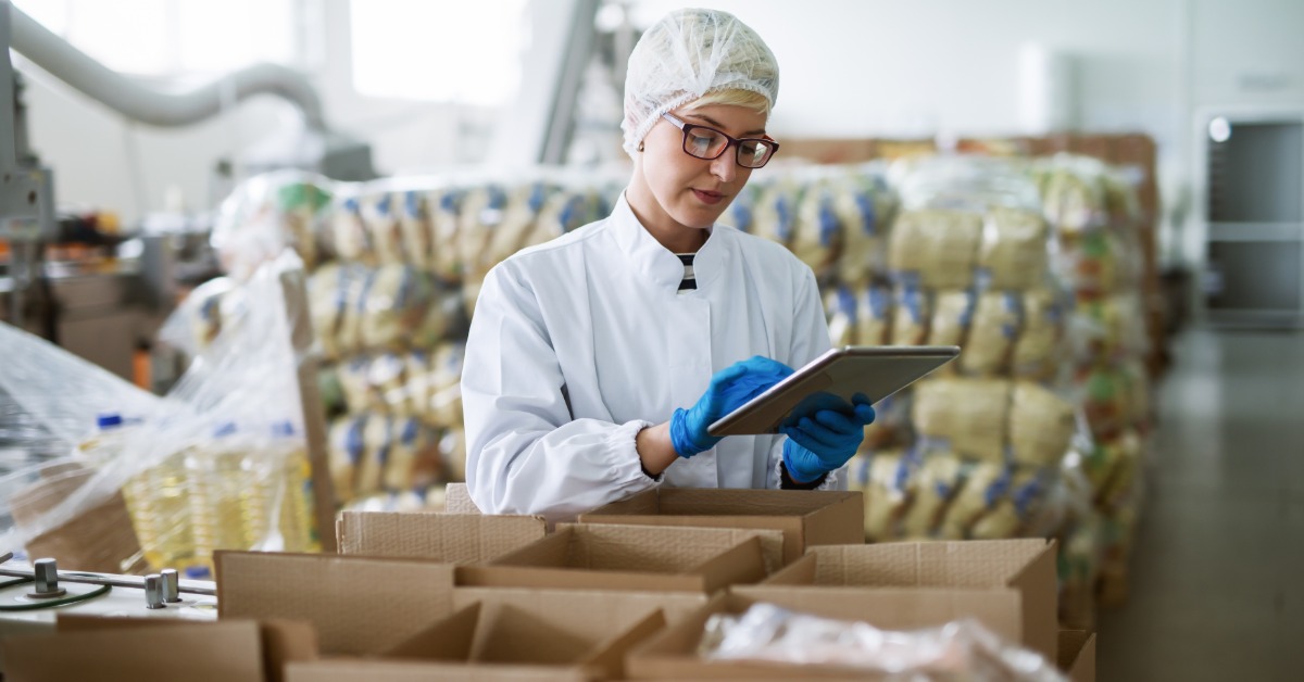 A woman dressed in white is in a food processing factory. She is holding a clipboard and is looking at it.