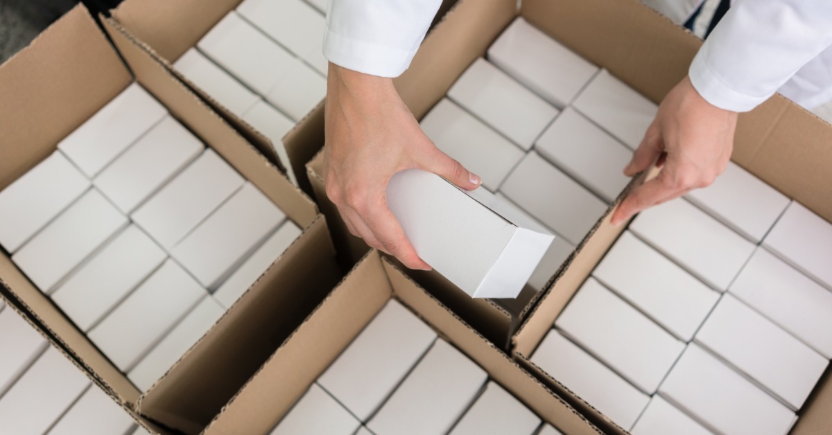 A person in a white coat is placing white boxes inside a larger cardboard box. There are other boxes on the same surface.