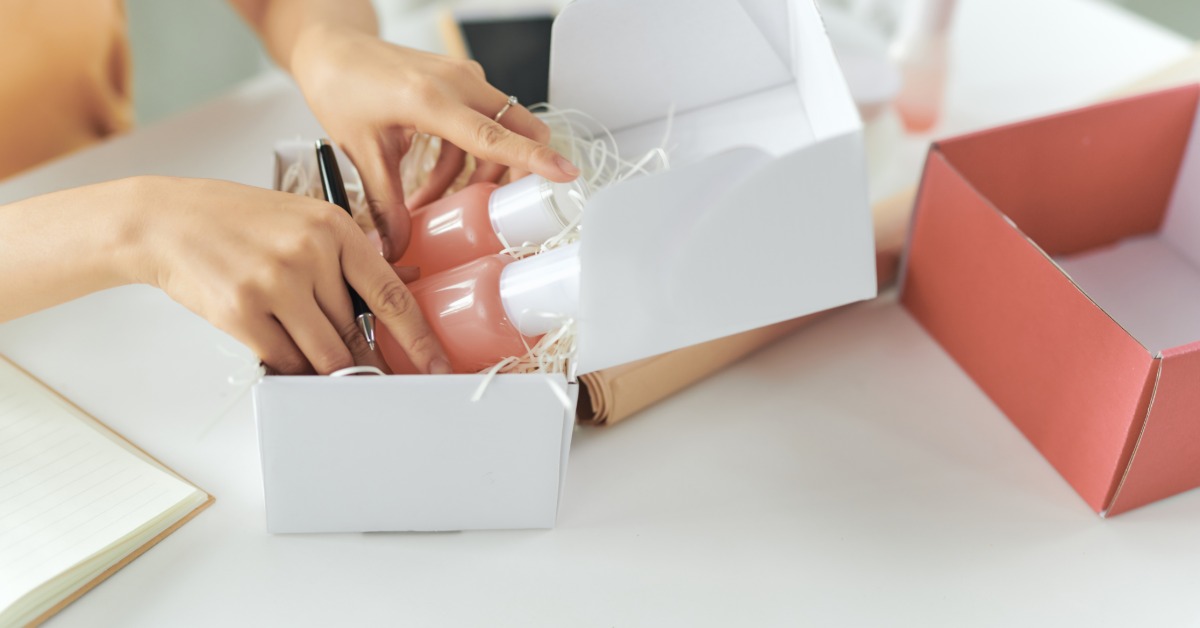 A woman is placing cosmetic supplies inside a box. The box has stuffing in it and the woman is holding a pen.