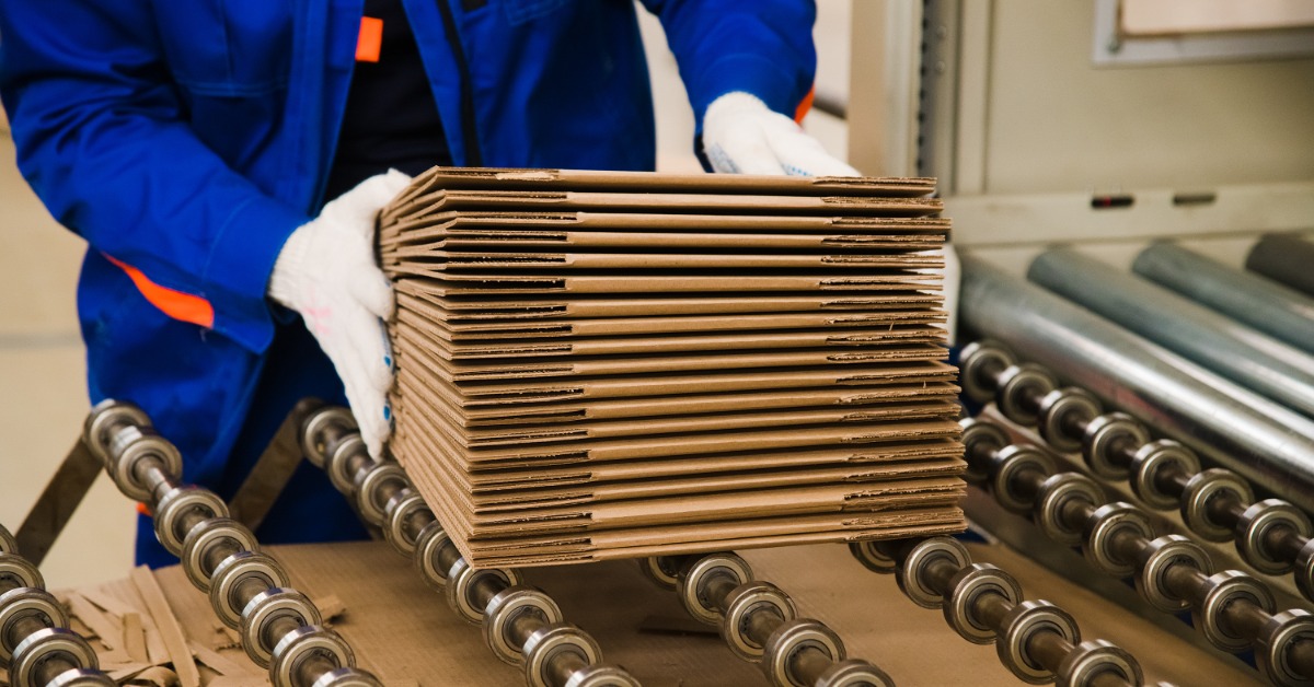 A person in a blue jumpsuit is holding a stack of unfolded cardboard pieces in their hands.
