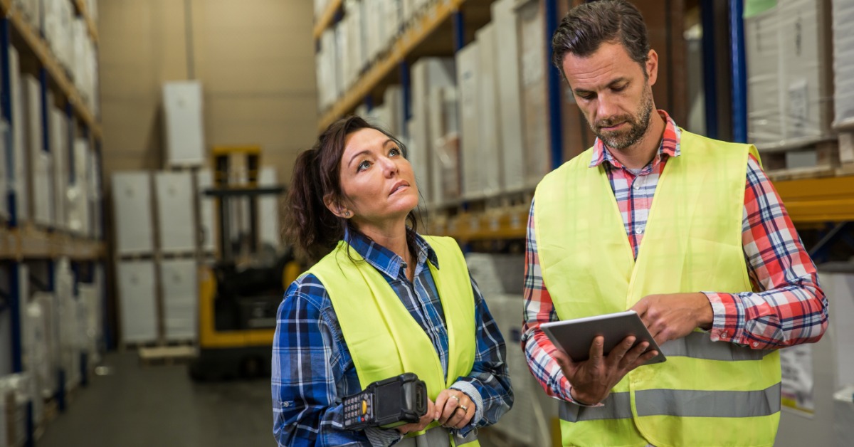 A man and a woman, both a whom are wearing flannel shirts and high-visibility vests, are in a warehouse area.