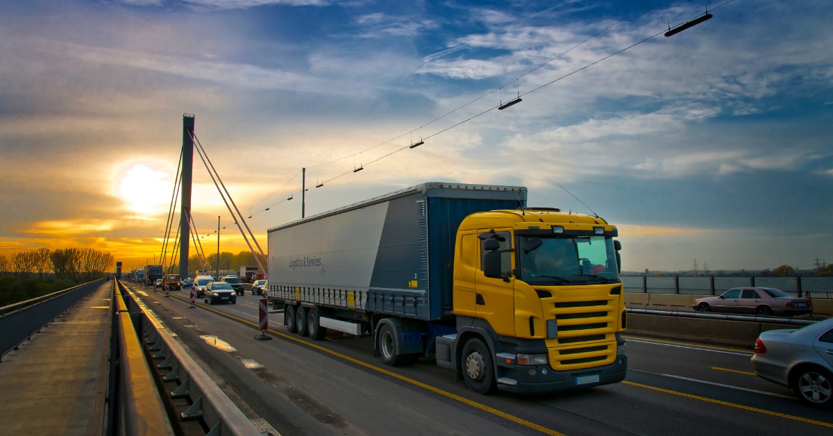 A truck is driving down a road. The sun is low in the sky in the background, and there are other cars on the road.