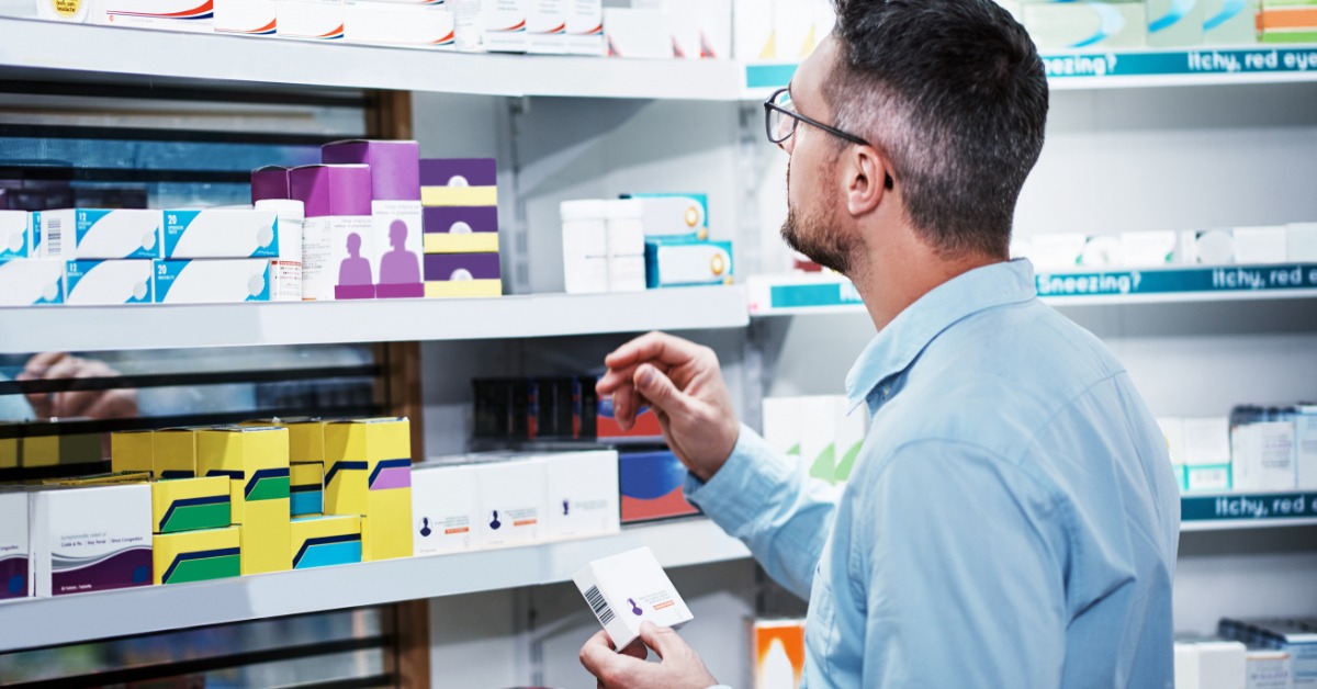 A man with glasses is looking at shelves filled with medicine boxes. The boxes range in color and size.