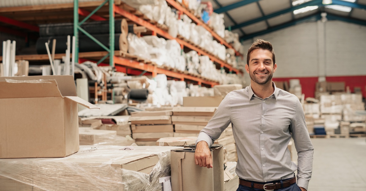 A man stands in a warehouse. There are multiple pallet racks in the background holding various materials.