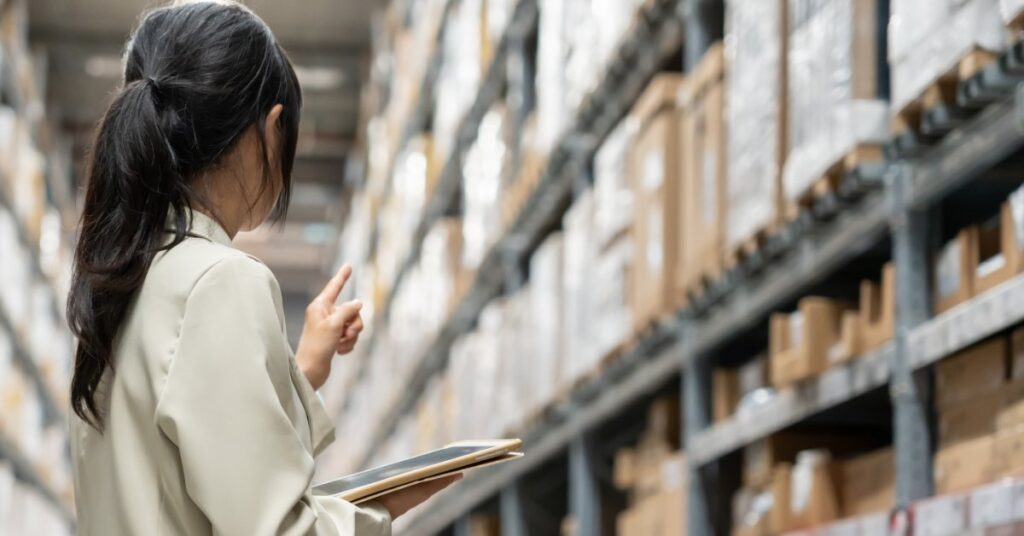 A woman is standing inside a warehouse and is pointing toward a wall of pallet racks. The racks all have loaded pallets.