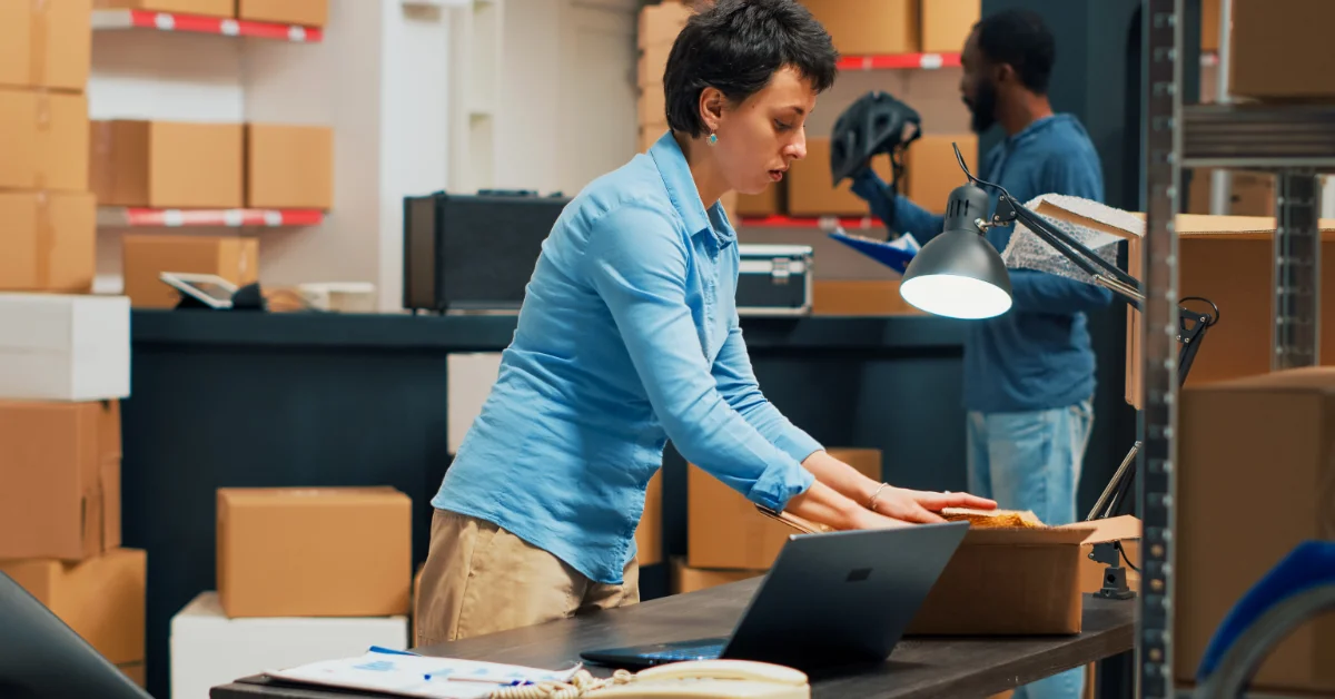 A woman with short hair is standing behind a desk. She is handling a box that is sitting on her desk.
