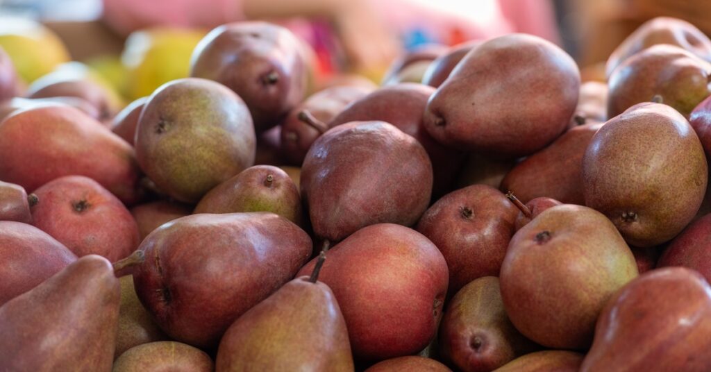 There are many red pairs in a pile. There are other pieces of produce in the background that are blurry.