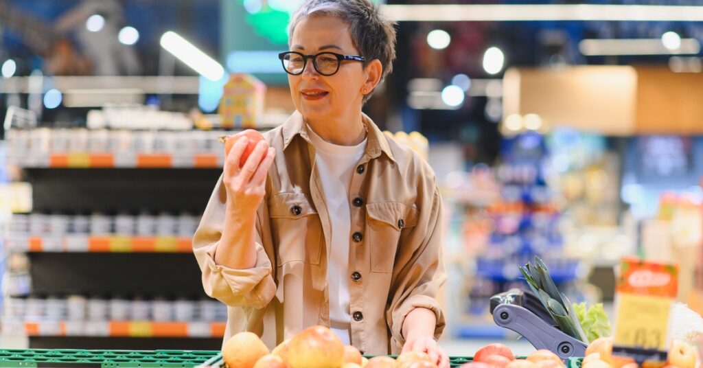 An older woman in a jacket it holding a piece of produce in her hand as she inspects it. She’s in a grocery store.