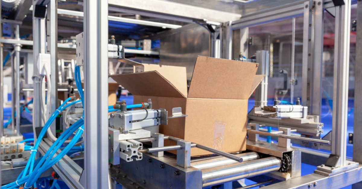 A cardboard box is in the center of an industrial machine. The machine has a blue base, and there are windows in the background.