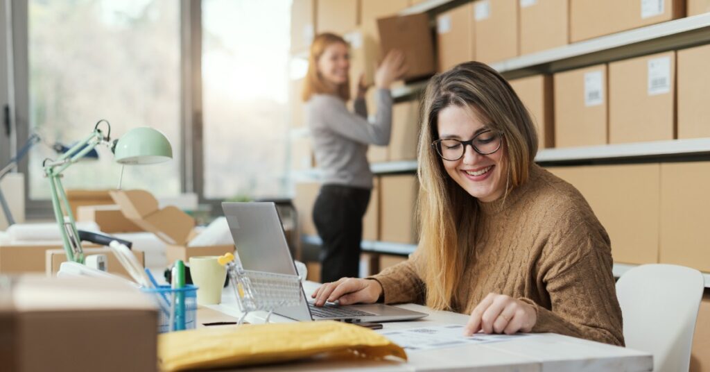 Two women are in a room. There is a wall of cardboard boxes, and one of the women is using a laptop.