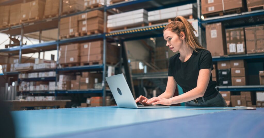 A woman is using a laptop in a warehouse environment. There are pallet racks with cardboard boxes on them.